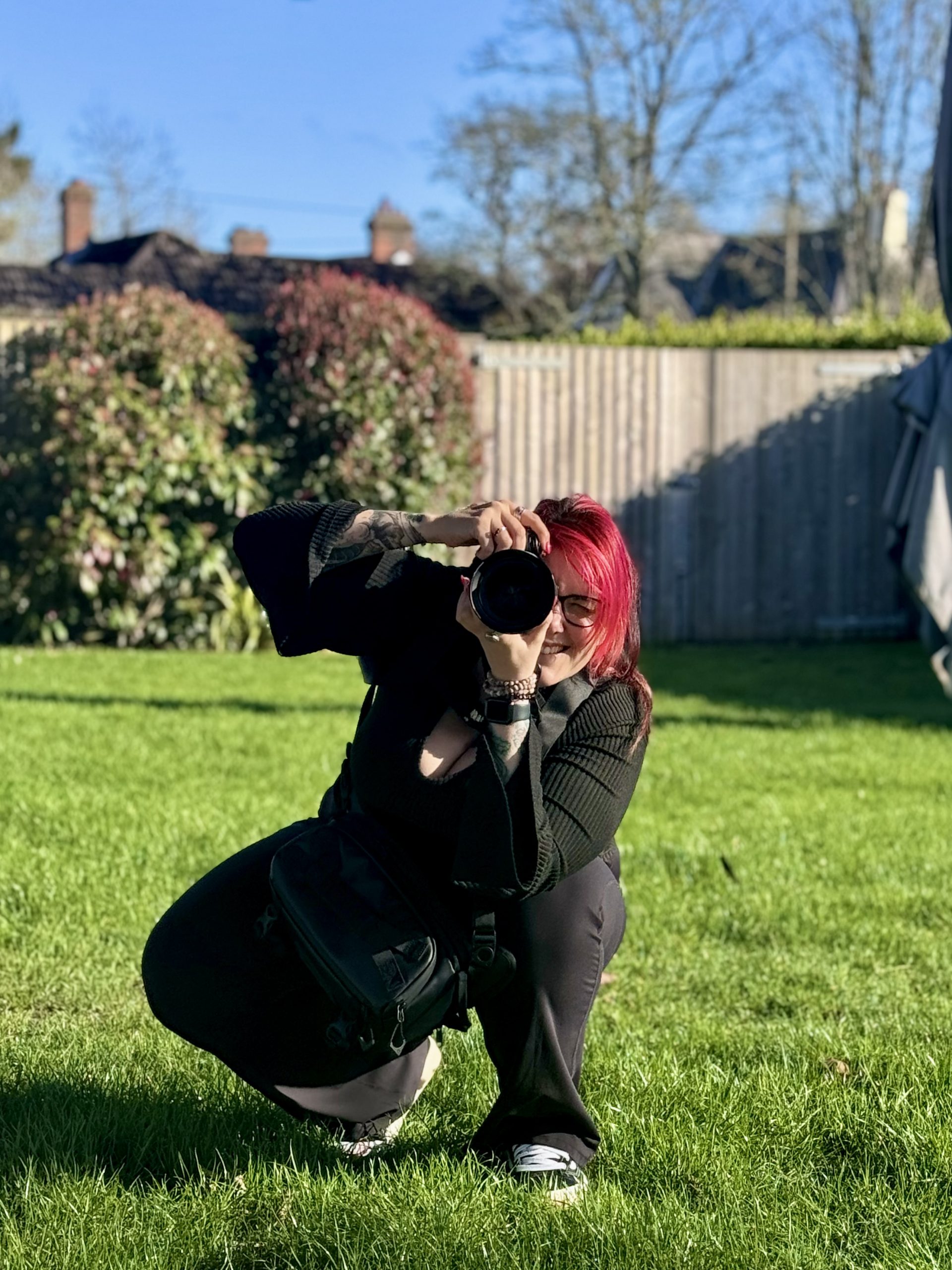 Becky Bryant Brand Photographer is at the Balmer Lawn Hotel, Hampshire. This is a photo of her squatting down, looking through her camera, ready to take a photo on a green grassy background with a blue sky